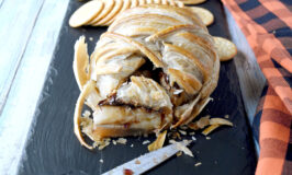 Oozing Mummy Baked Brie is topped with dried cranberries simmered in orange juice and dark chocolate chips. It’s a sweet and savory treat for an appetizer or dessert and super simple to make. #HalloweenTreatsWeek #bakedbrie